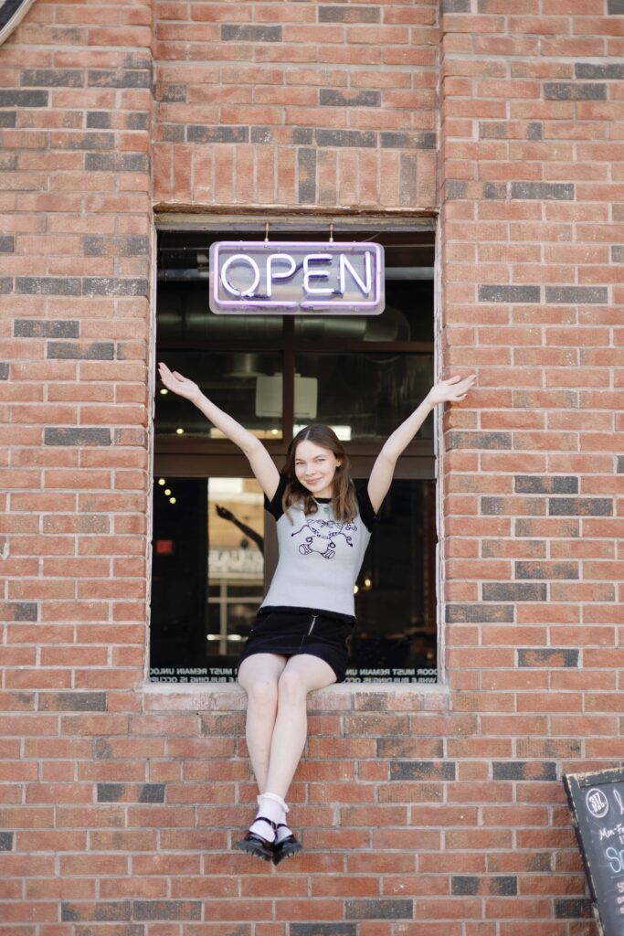 Senior girl posing with arms up sitting on brick wall in Broad Ripple in Indianapolis.